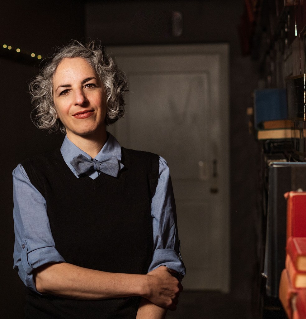 Portrait of me, wearing a light blue collared shirt and matching bow tie with a black sweater vest. I have gray hair and dark eyes and eyebrows. I'm standing in a dark hallway with one arm across my belly gripping my other arm, which is hanging straight down. To my right are some books on a shelf.